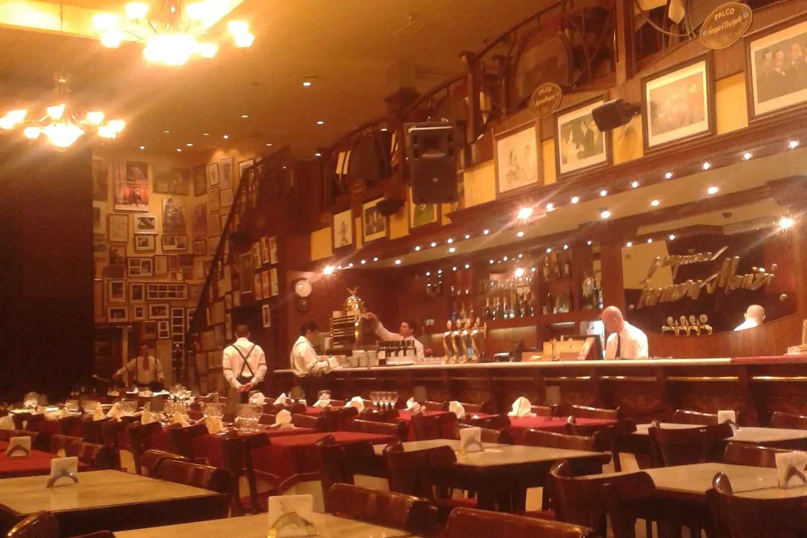 The dining hall of Esquina Homero Manzi before service, with marble-topped tables, red cloths, an upper gallery of boxes hung with framed pictures and a long bar